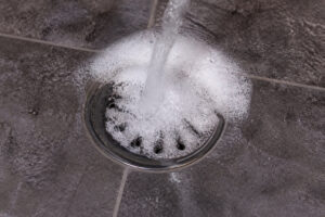 Eau savonneuse s'écoulant dans une bonde de douche en métal sur un sol en carrelage gris.