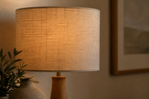 Abat-jour cylindrique en tissu beige diffusant une lumière chaleureuse sur un pied de lampe en bois clair.