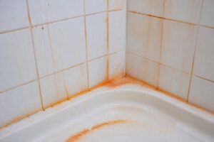 Gros plan sur un coin de douche en carrelage blanc avec des traînées de rouille orange sur les joints et le receveur.