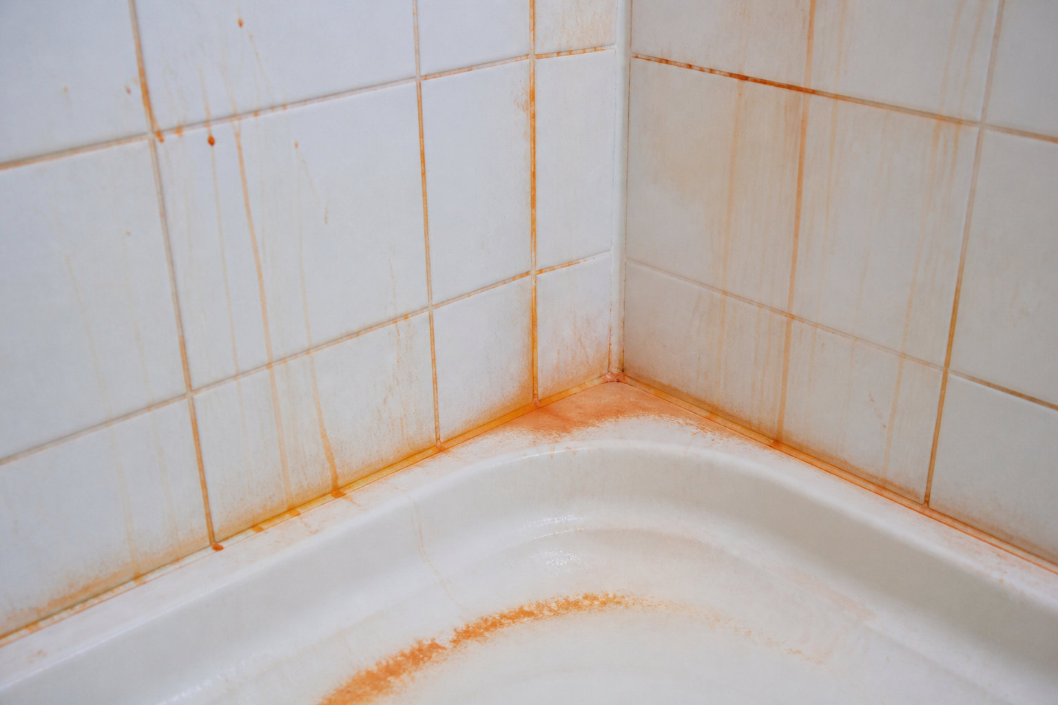 Gros plan sur un coin de douche en carrelage blanc avec des traînées de rouille orange sur les joints et le receveur.